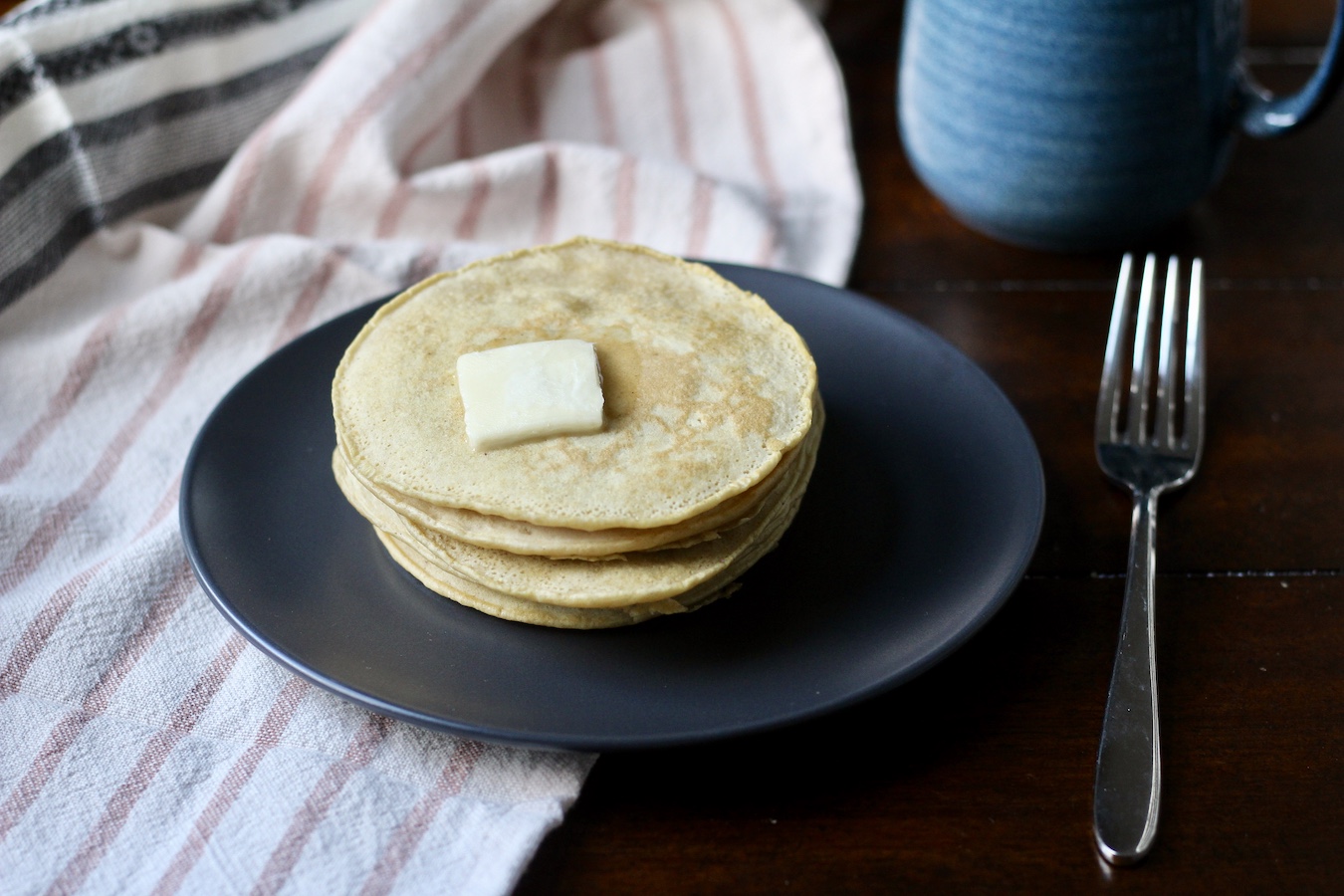 A stack of fresh buttermilk pancakes with a small square of vegan butter on top, on a blue plate on a white and pink striped dish towel wit a silver fork to the right and a blue coffee mug in the back right on a wooden table.