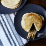 A blue plate with a stack of buttermilk pancakes with maple syrup on top and a triangle of pancakes cut out and on a silver fork in front with a large blue plate of buttermilk pancakes in the back on a white and blue striped dish towel on a wooden table.