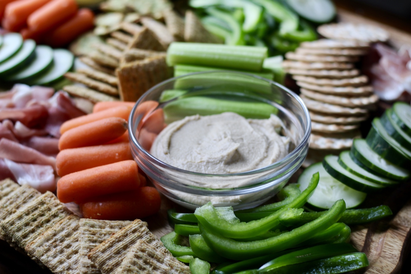 Image of hummus on a charcuterie board with veggies and crackers around it. 