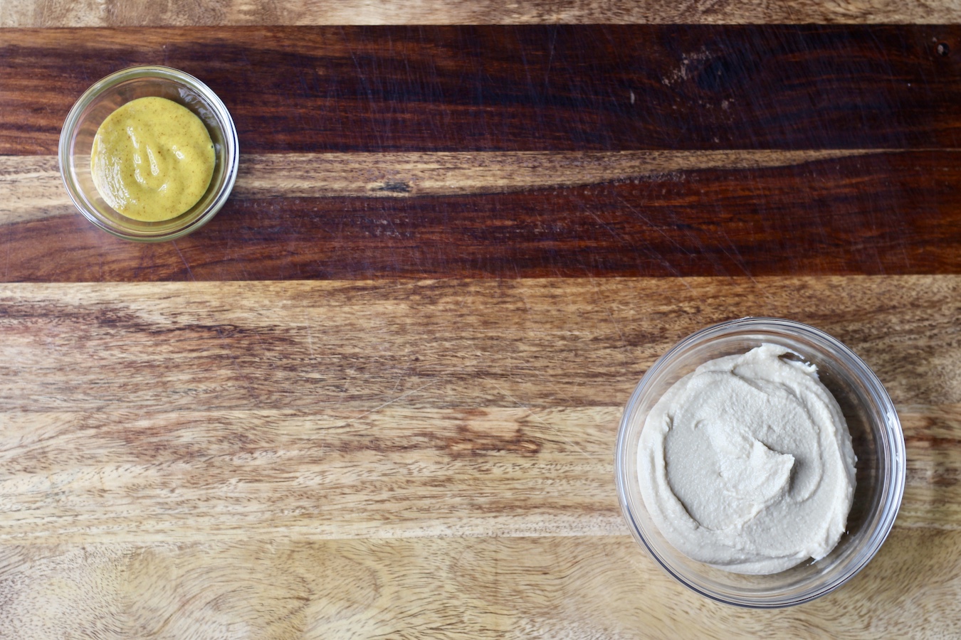 Wooden cutting board with honey mustard and hummus in small glass bowls.