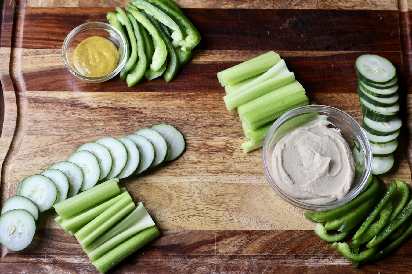 Wooden cutting board with honey mustard and hummus in small glass bowls and celery, cucumber, and bell pepper next to the bowls.