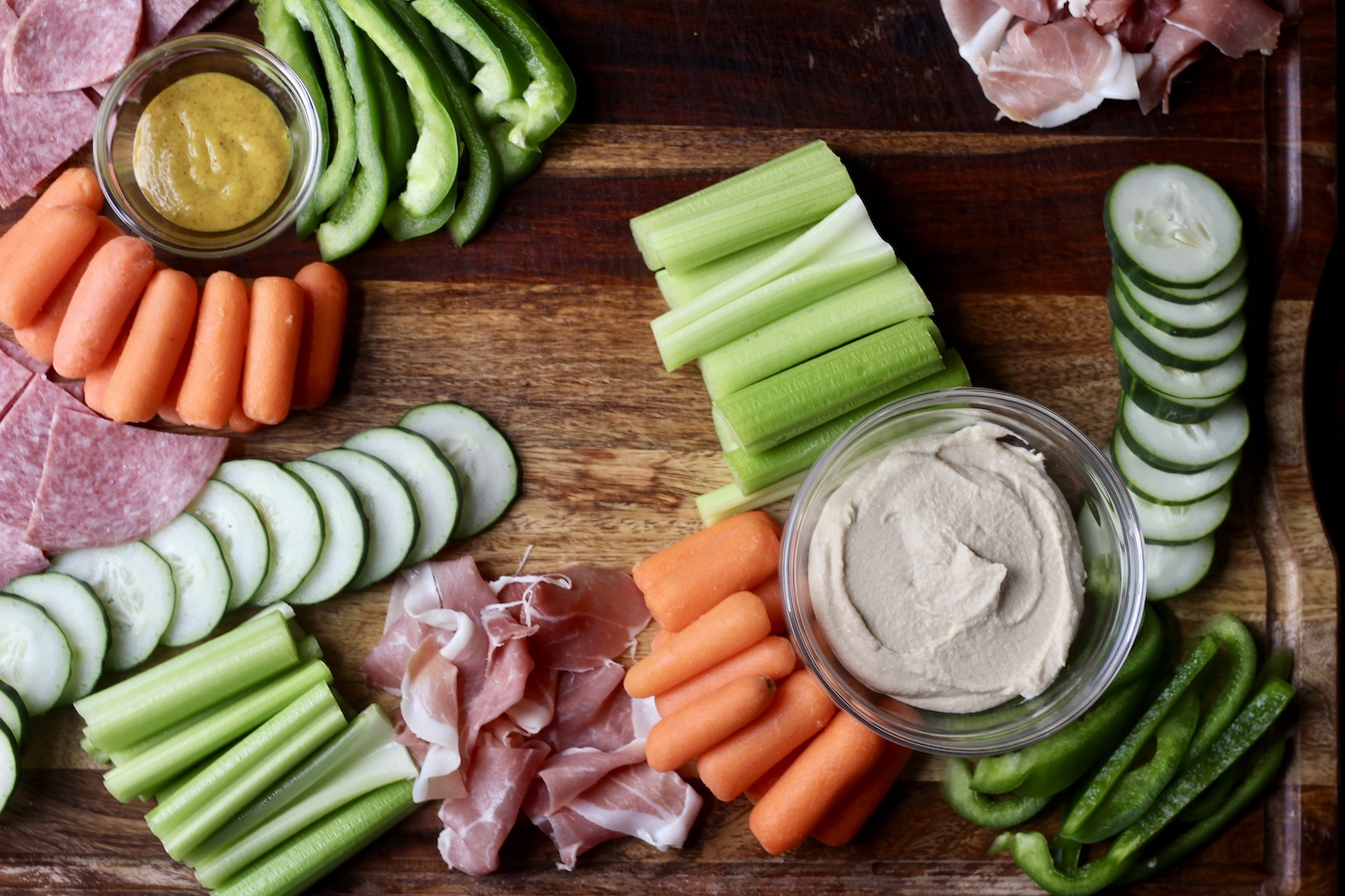 Wooden cutting board with honey mustard and hummus in small glass bowls and celery, cucumber, and bell pepper next to the bowls with carrots, salami, and prosciutto in between.