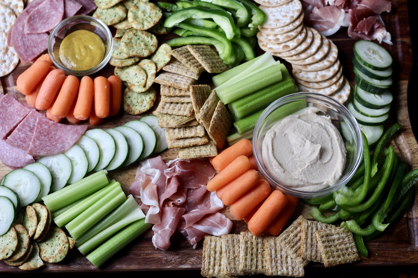 Finished charcuterie board with celery, carrots, cucumber, bell pepper, salami, prosciutto, crackers, hummus, and honey mustard.