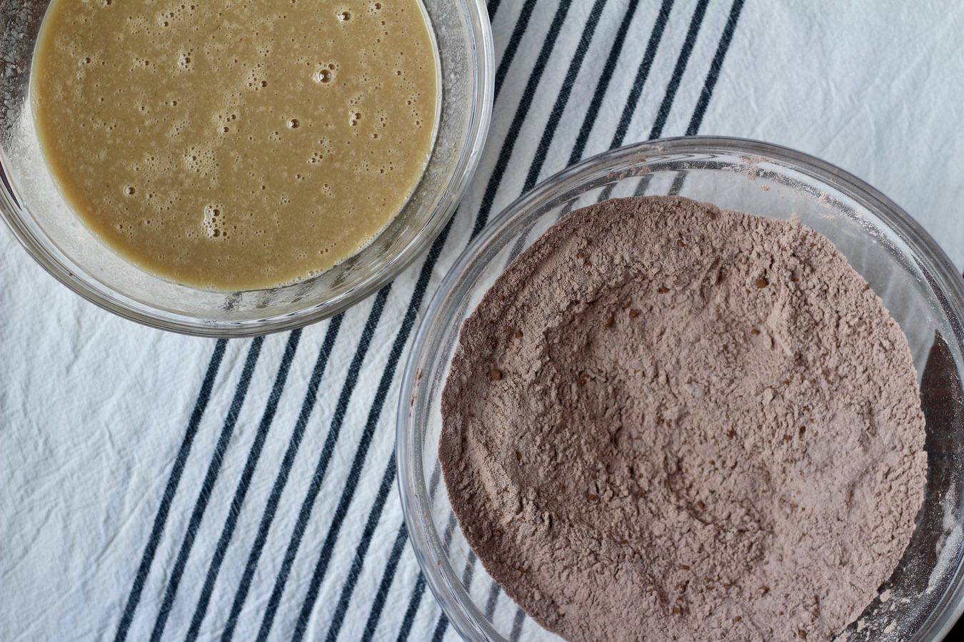 One bowl of liquid ingredients and one bowl of dry ingredients on a white and blue striped dish towel.