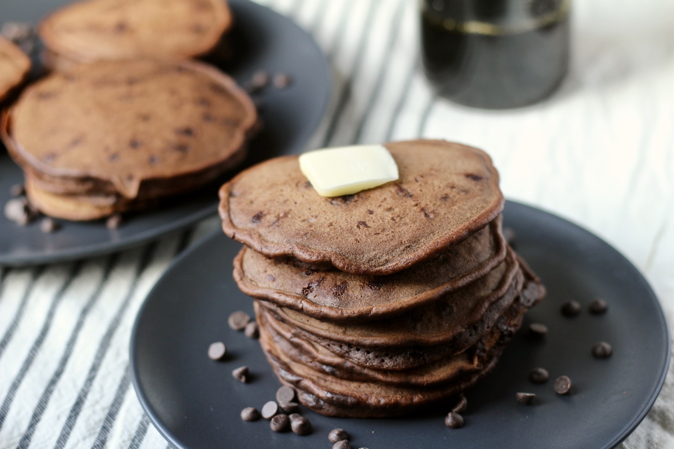 Two blue plates of double chocolate chip pancakes with a pad of butter on the front plate's stack on a white and blue striped dish towel with syrup in the back.