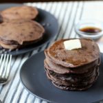 Two blue plates of double chocolate chip pancakes with a pad of butter on the front plate's stack on a white and blue striped dish towel with syrup in the back and forks to the left.