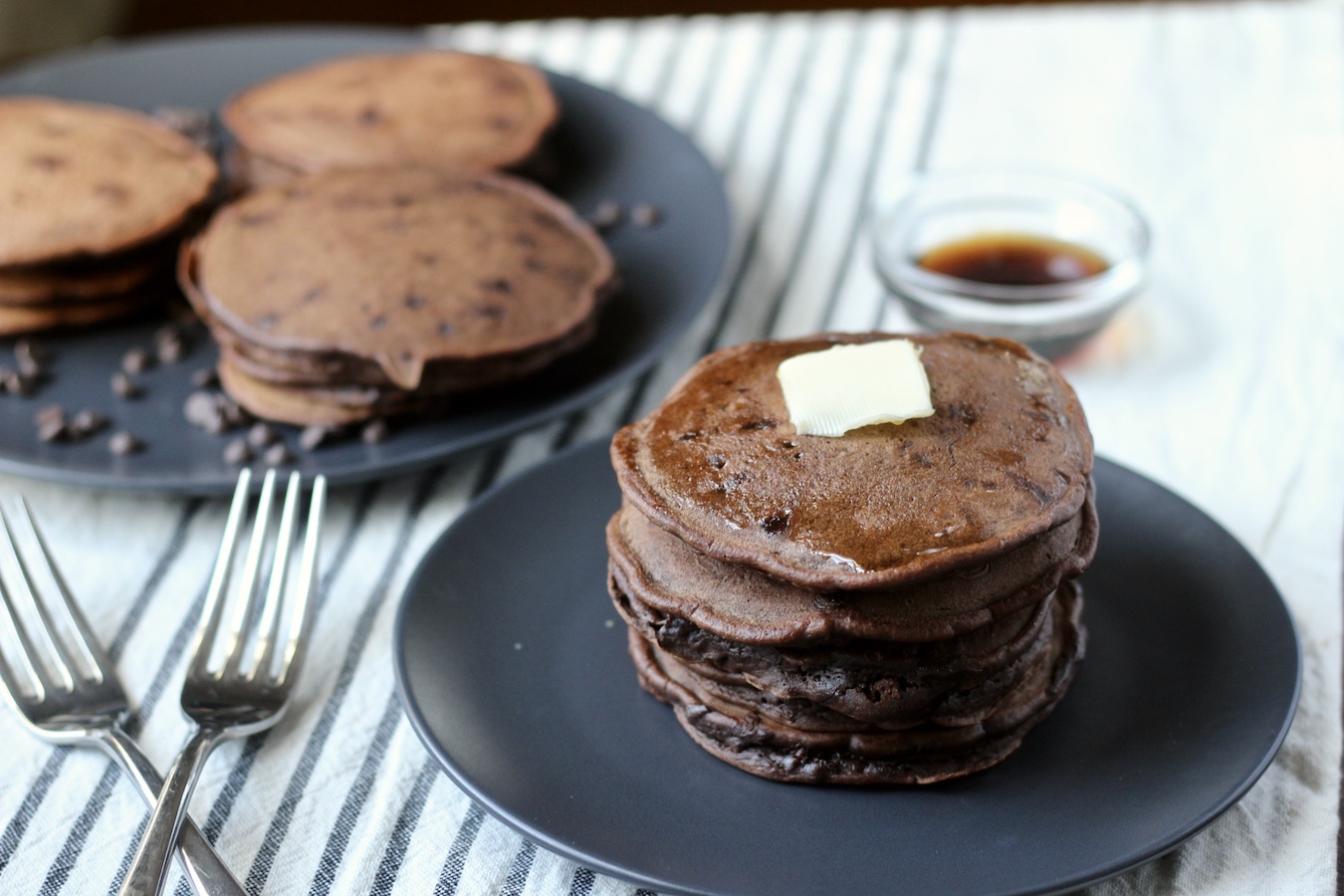 Two blue plates of double chocolate chip pancakes with a pad of butter on the front plate's stack on a white and blue striped dish towel with syrup in the back and forks to the left.
