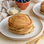 A close up of a white plate with a brown rim stacked with buttermilk pancakes with a small dish of vegan butter in the back and a gold fork in the front right.