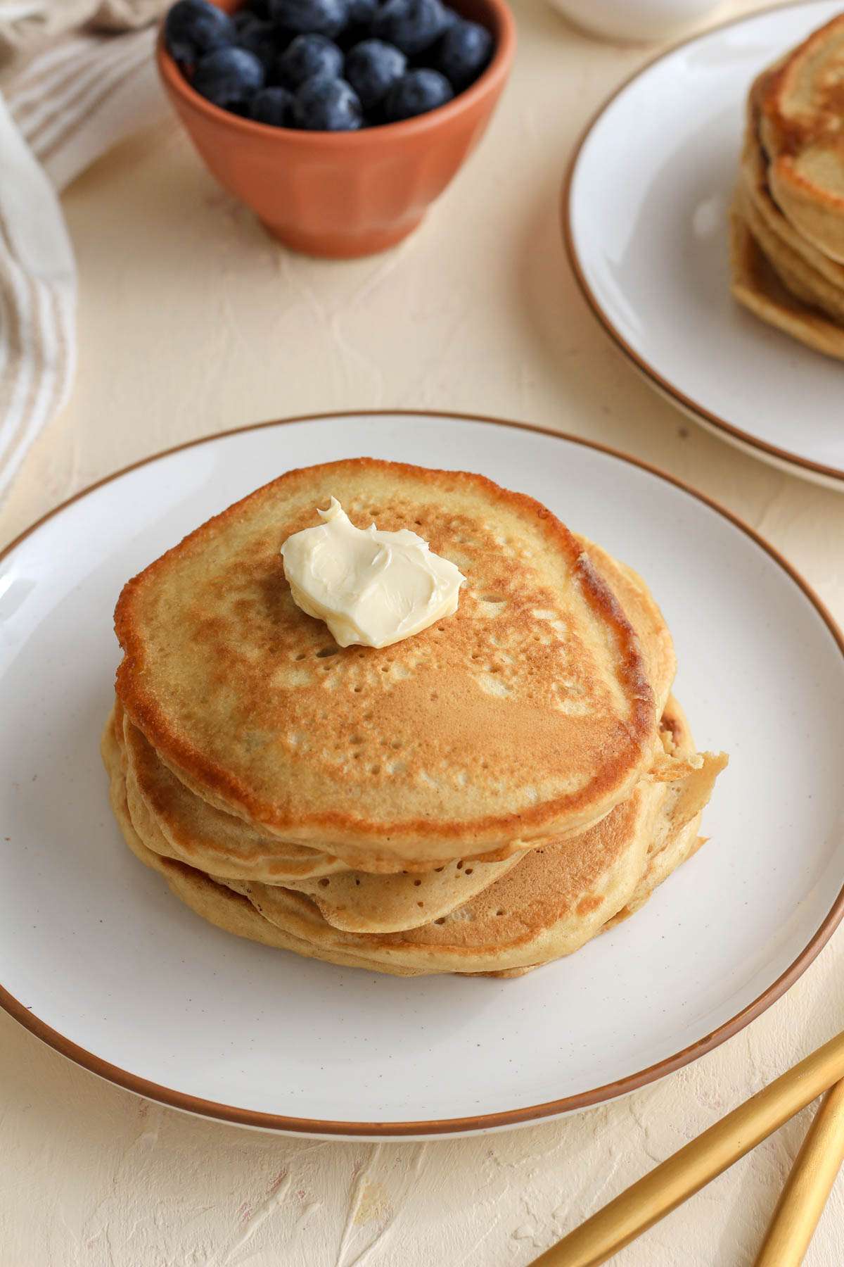 A white plate with a brown rim topped with buttermilk pancakes and a pad of vegan butter on top.