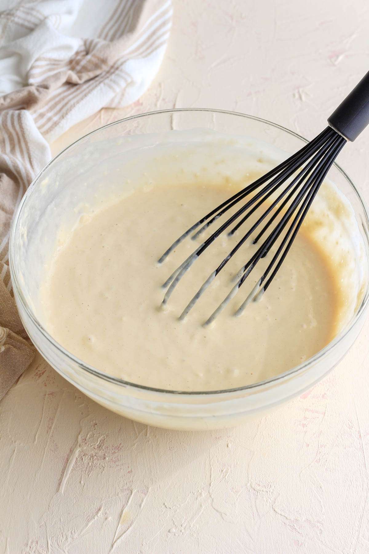 A glass mixing bowl with dairy-free buttermilk pancake batter mixed with a black whisk.