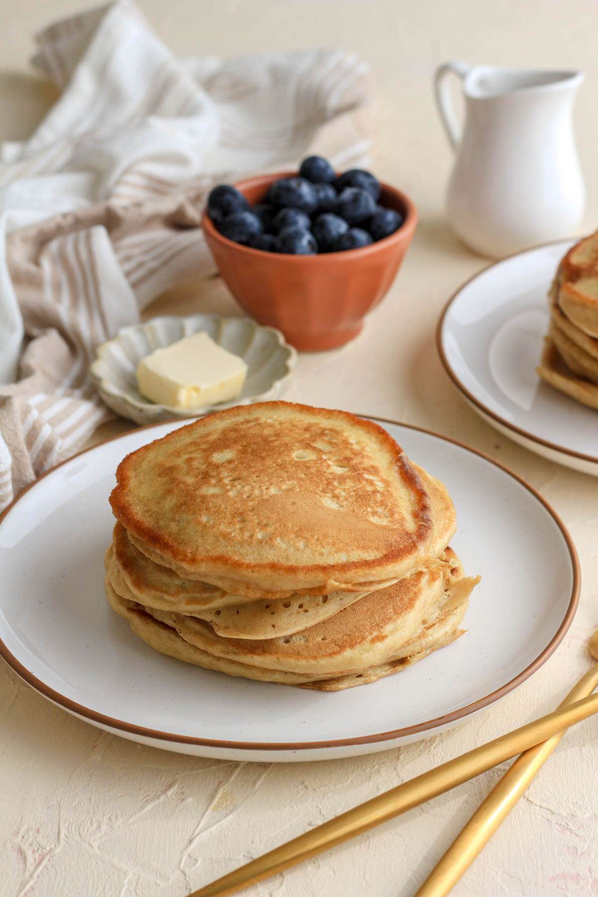 A white plate with brown rim topped with dairy free pancakes made with vegan buttermilk and a small dish with vegan butter and a small bowl of blueberries in the back.