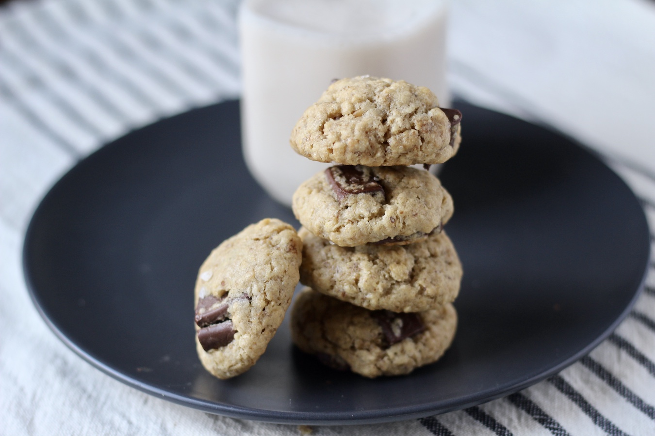 A small stack of lactation cookies with a bottle of almond milk in thee back on a blue plate on a white and blue striped dish towel.