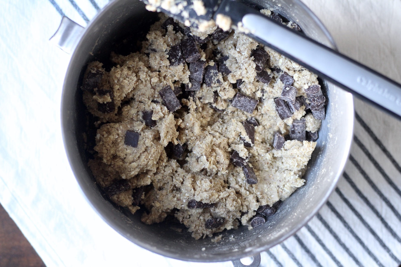 Raw cookie dough in a silver mixing bowl with a black spatula on a white and blue striped dish towel.