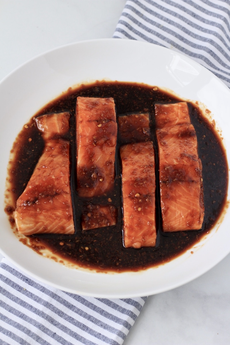 Salmon fillets marinating in a white bowl on a counter with a blue and white striped towel.