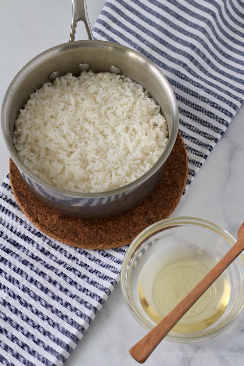 White long grain rice in a sauce pan with a small bowl of vinegar mix for the rice in the bottom right corner.