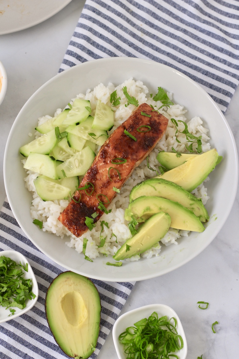 A white bowl with sticky rice topped with cucumbers, salmon, and avocado on a blue and white striped towel.