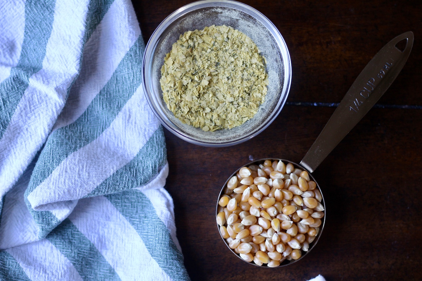 Popcorn kernels in a measuring cup with nutritional yeast seasoning in a bowl next to a green and white towel.