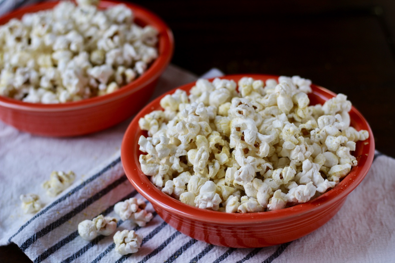 Two orange bowls of nooch popcorn on a white and blue dish towel with popcorn on the table.