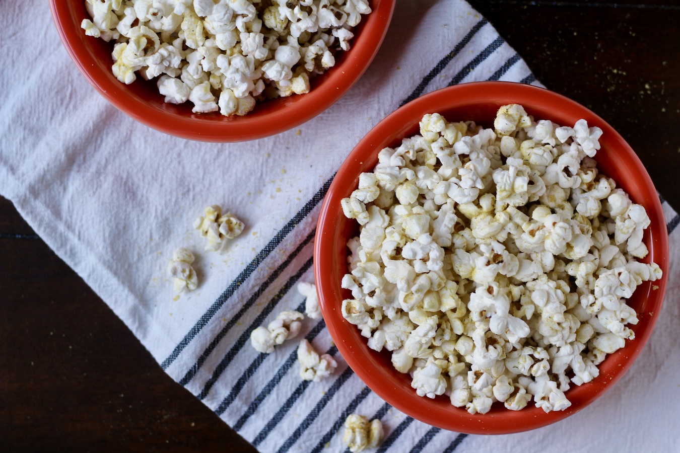 Two orange bowls of stovetop popcorn with nooch and herbs on a white and blue striped towel with a few pieces of popcorn between the two.