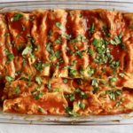 A glass pyrex dish with a dozen baked stuffed turkey enchiladas with enchilada sauce and chopped cilantro on a white and blue striped dish towel.