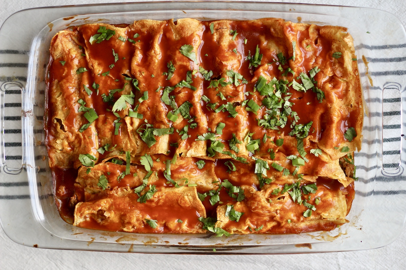 A glass pyrex dish with a dozen baked stuffed turkey enchiladas with enchilada sauce and chopped cilantro on a white and blue striped dish towel.