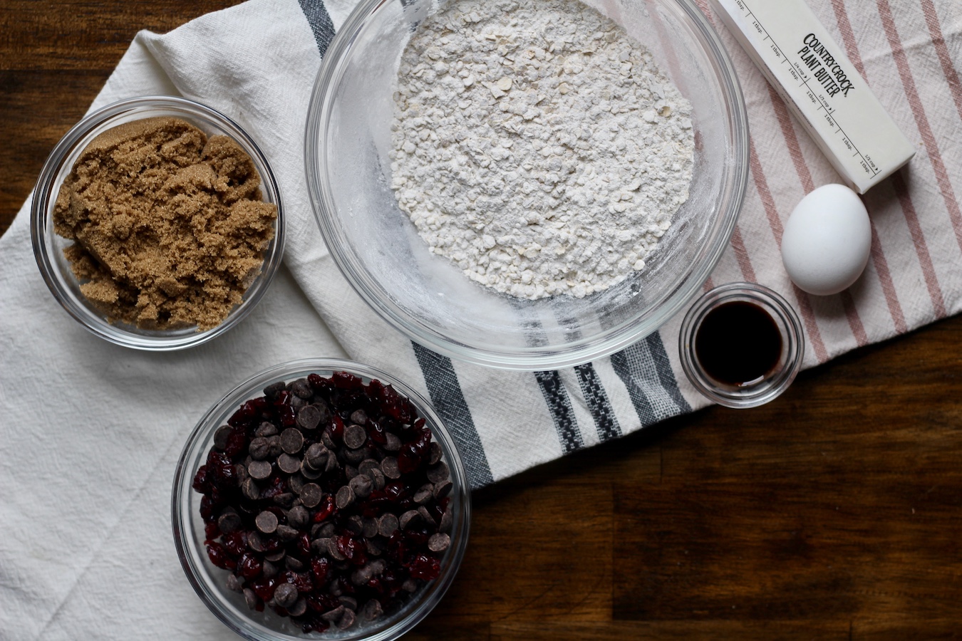 Ingredients for oatmeal craisin cookies in glass bowls with pink, blue and white striped dish towel on a wooden table.