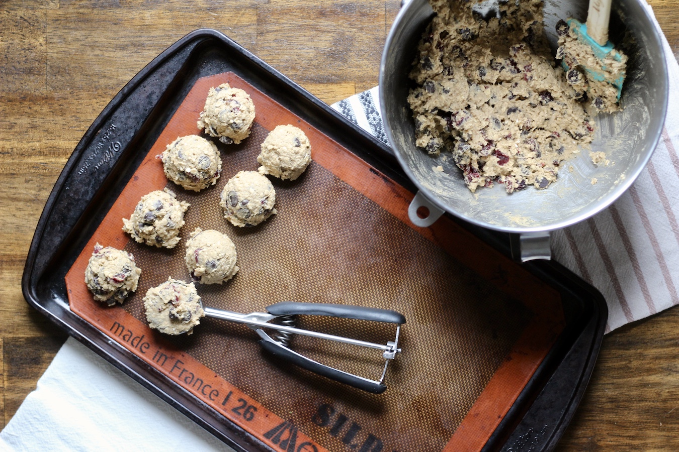 A mixing bowl of cookie dough with a cookie sheet, that has 8 balls of dough with a cookie scoop on a blue, white and pink dish towel on a wooden table.