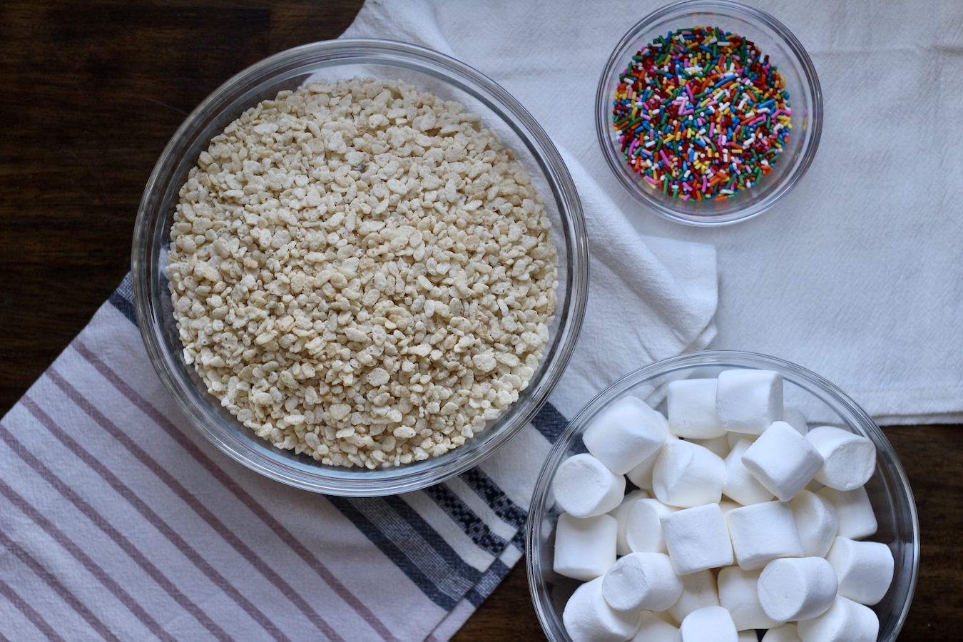 Three glass bowls with ingredients for funfetti rice crispy treats. One has marshmallows, one has rice crispy cereal, and one had funfetti sprinkles.
