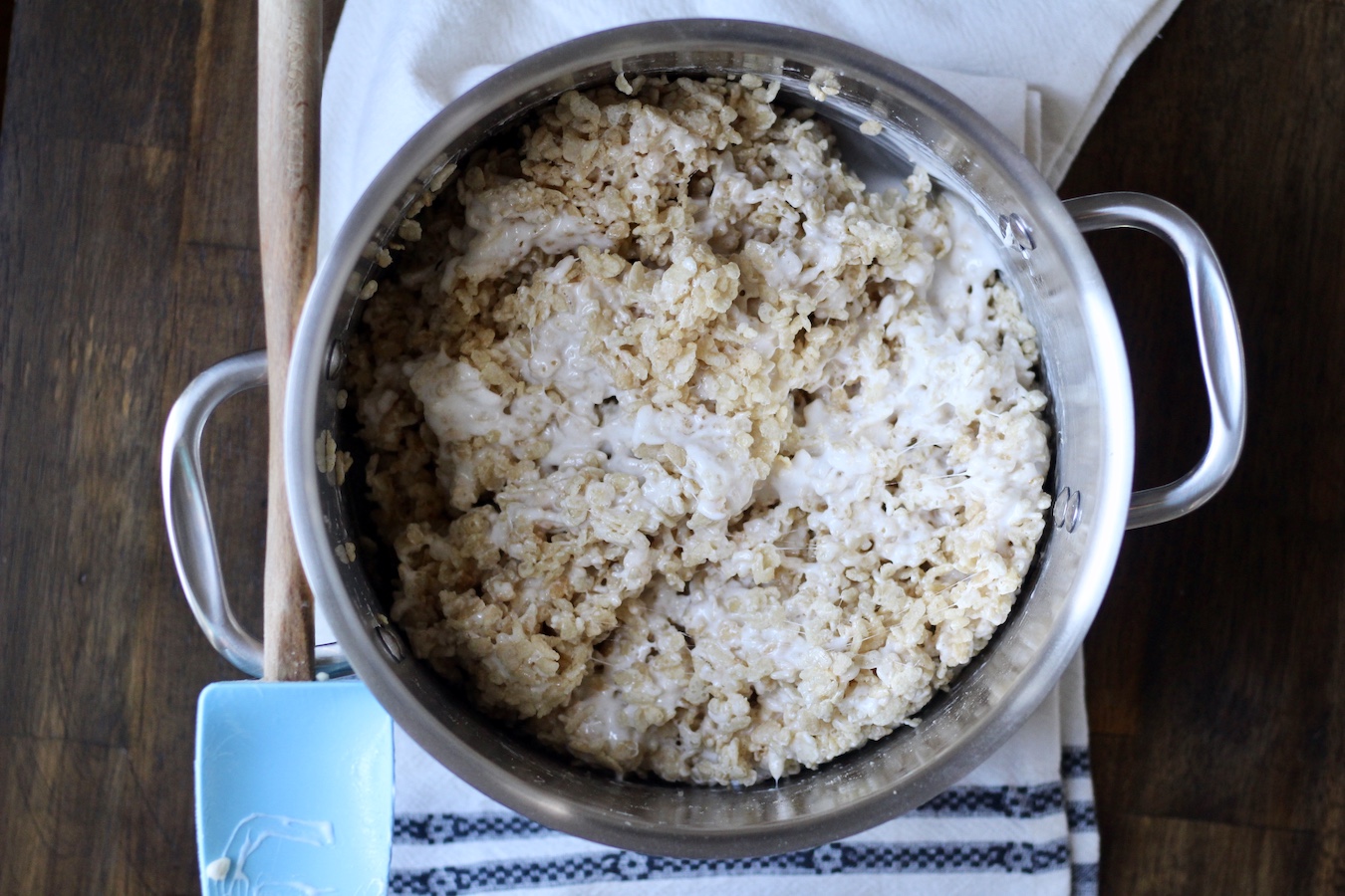 Rice crispy cereal covered in the marshmallow mixture in a stainless steel pot with a rubber spatula on a white dishtowel on a wooden table.