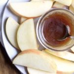 A white plate with apple slices and a small jar of salted caramel sauce with a spoon on a wooden table with a blue and white dish towel.