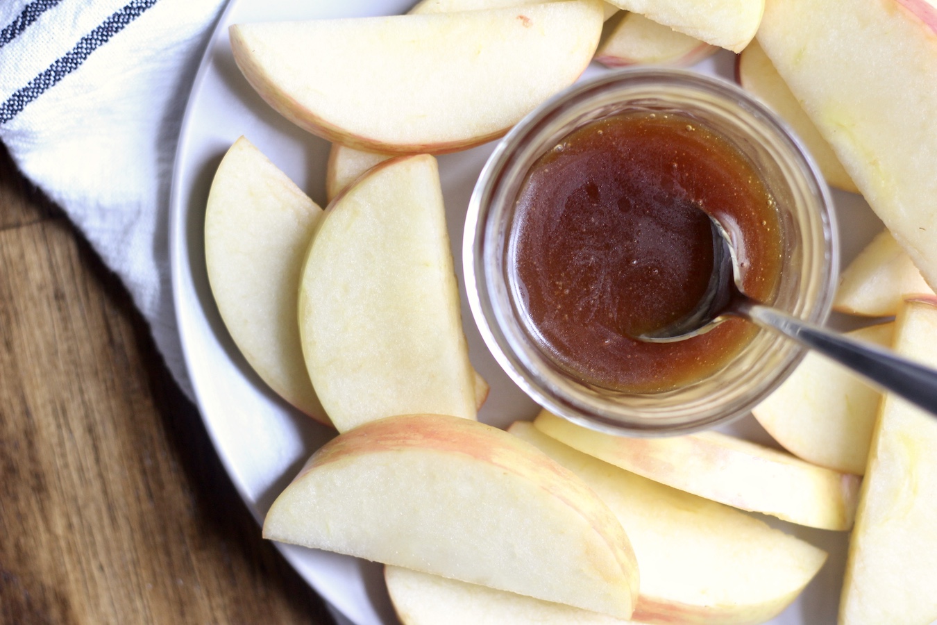 A white plate with apple slices and a small jar of salted caramel sauce with a spoon on a wooden table with a blue and white dish towel.