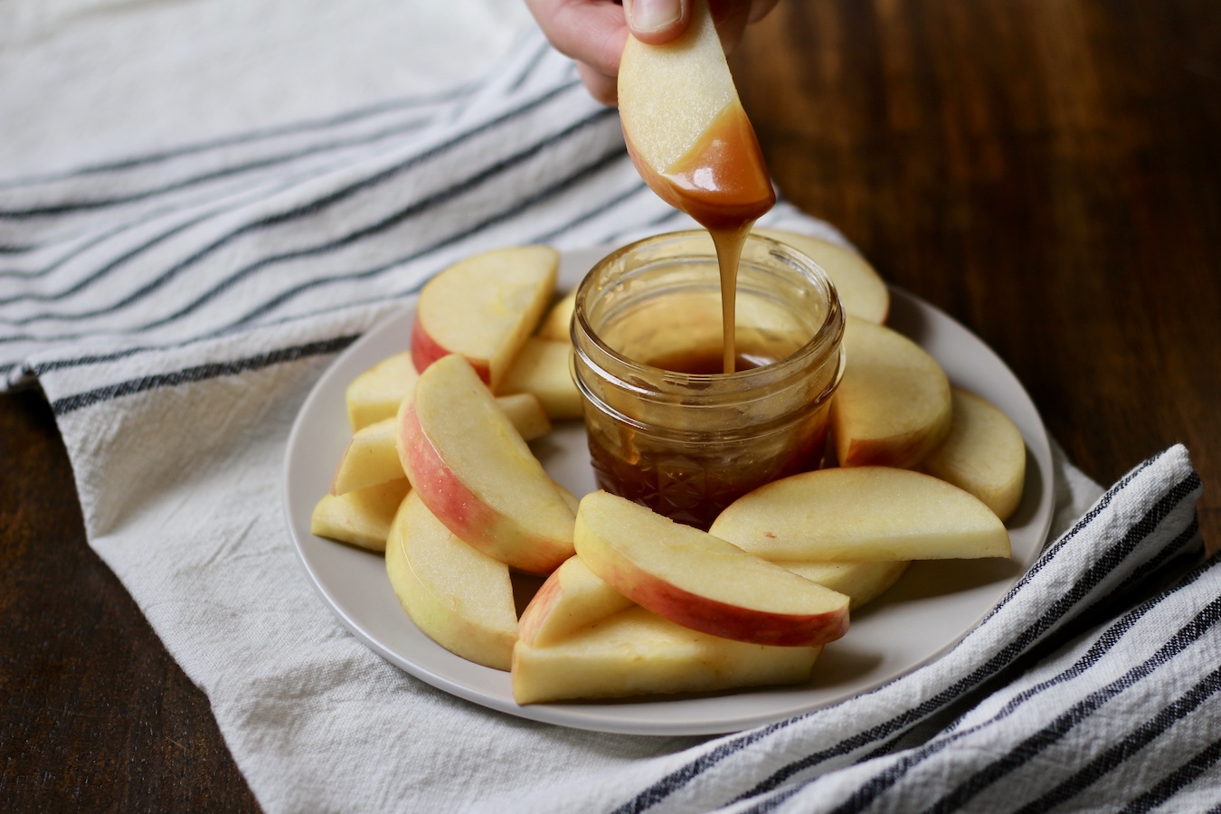 A white plate with apple slices and a small jar of salted caramel sauce with a hand dipping an apple in the sauce on a wooden table with a blue and white dish towel.