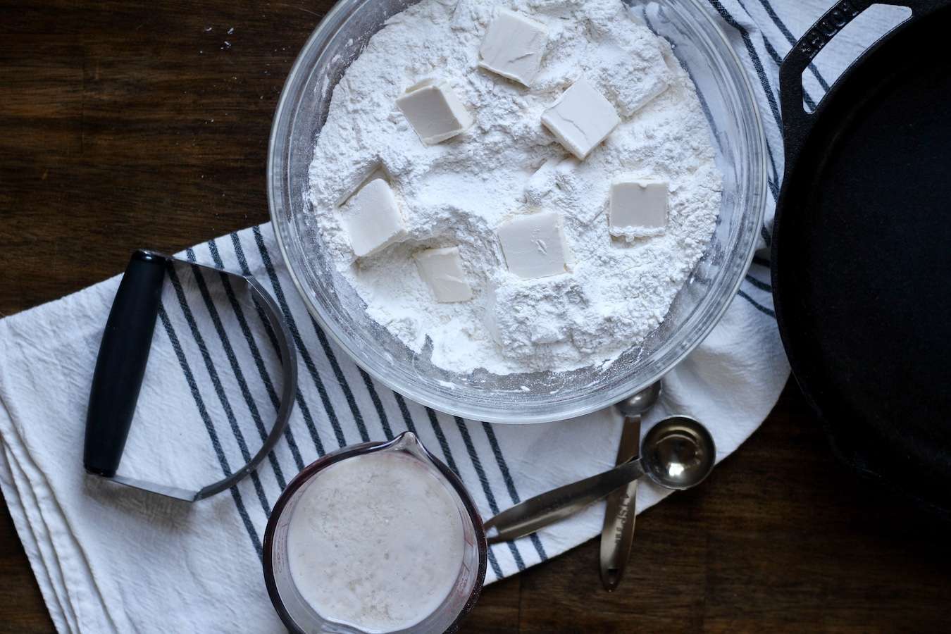 Glass bowls with flour and butter mixture, non-dairy buttermilk, measuring spoons, and a pastry blender on a white and blue towel on a wooden counter.