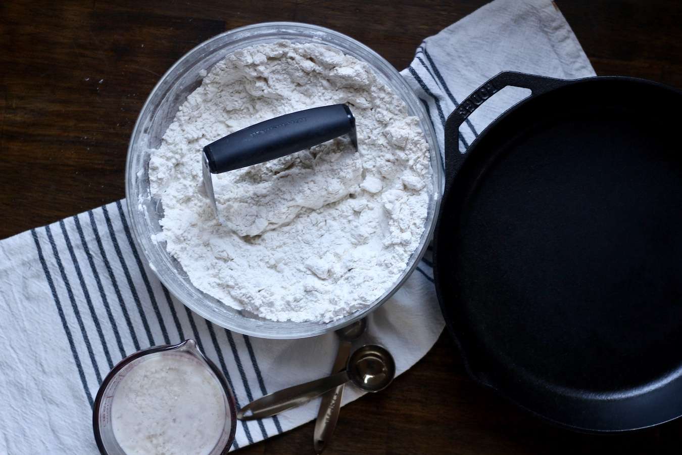 Pastry blender in the flour butter mixture bowl with a cup of non-daiy buttermilk next to a cast iron skillet on a white and blue towel on a wooden table.
