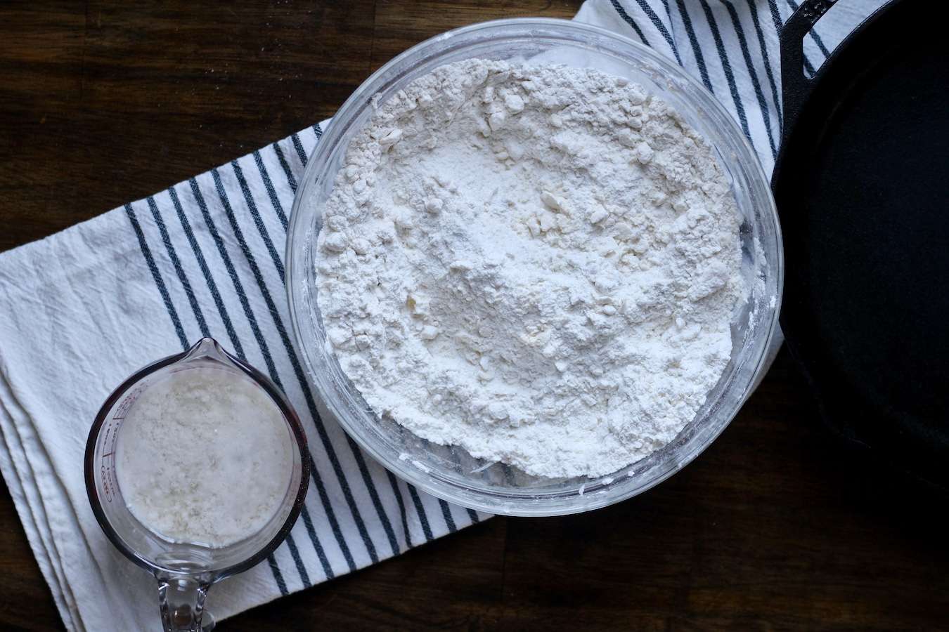 A bowl of the flour butter mixture with a cup of buttermilk to the side on a white and blue dish towel on a wooden table.