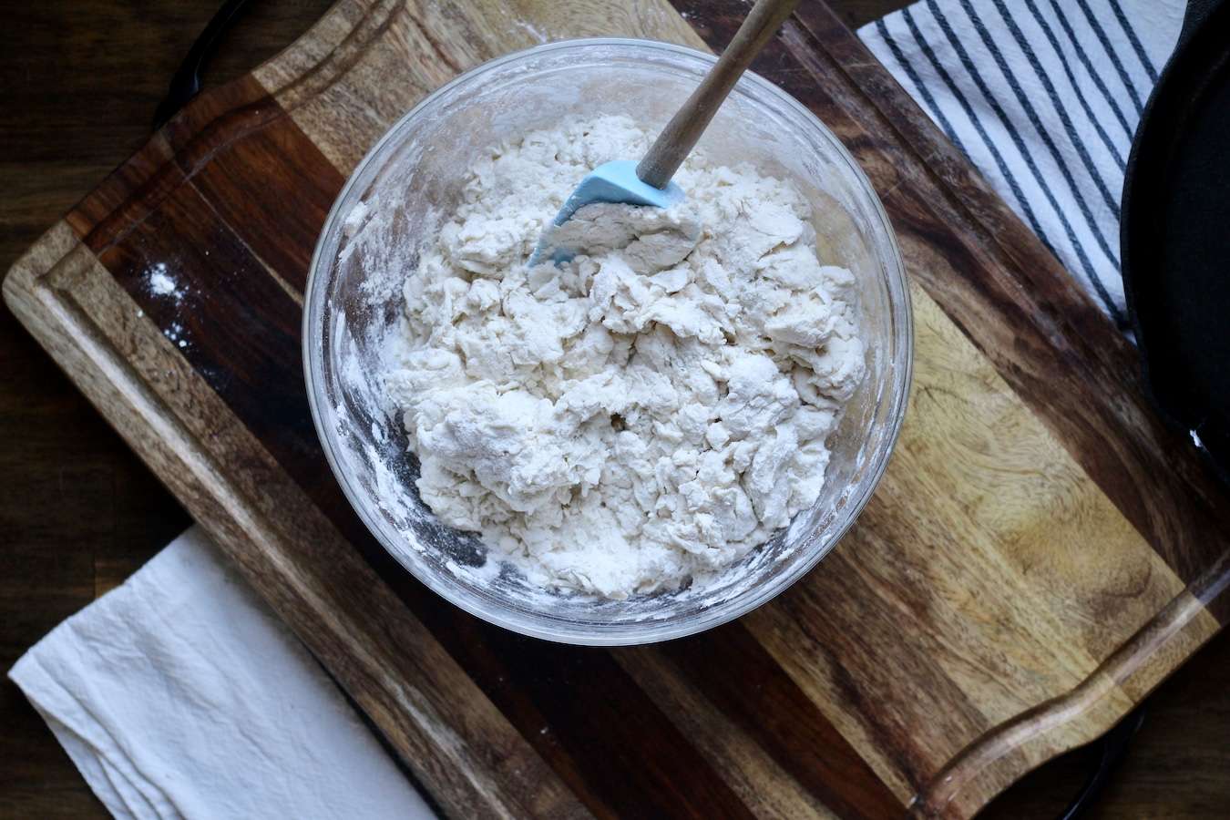 Buttermilk biscuit dough in a glass bowl with a spatula on a wooden cutting board.