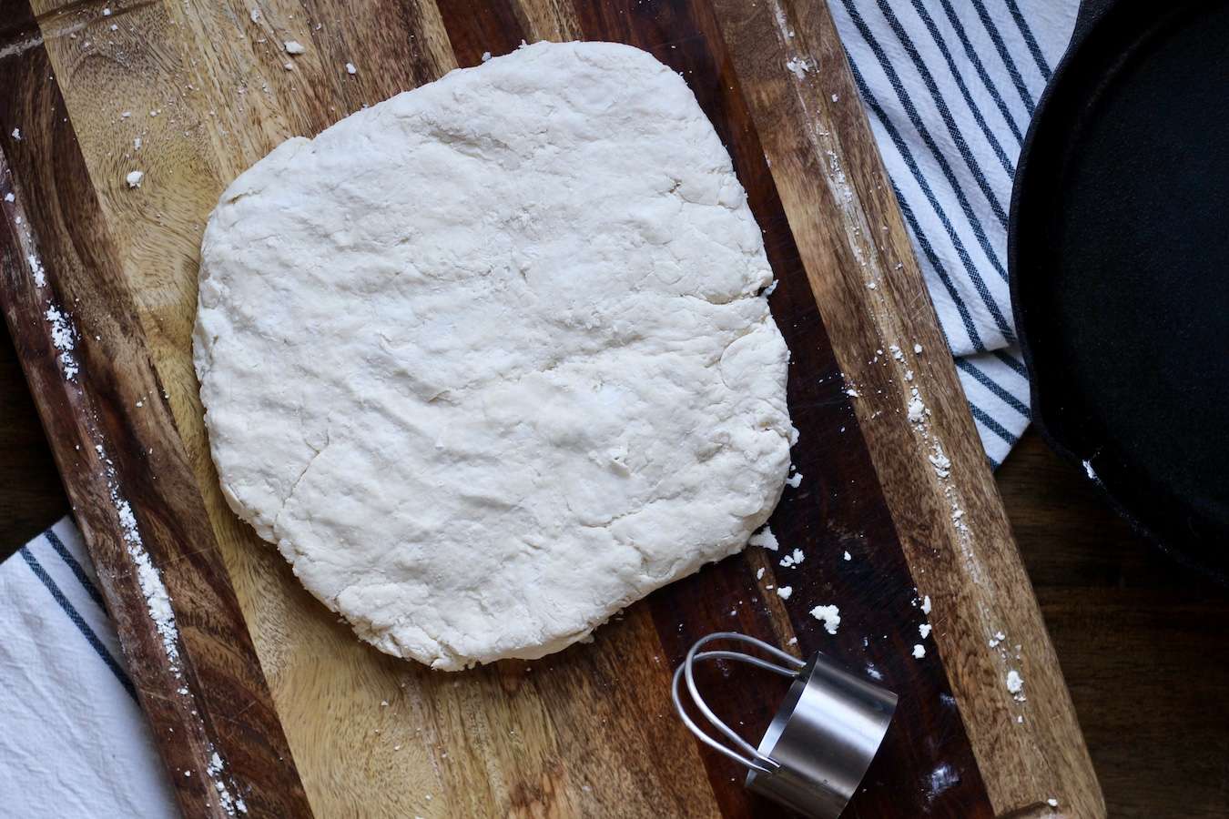 Biscuit dough on a cutting board with a biscuit cutter and the cast iron skillet.