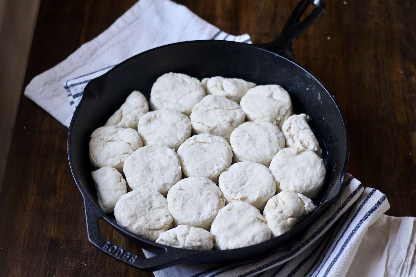 A cast iron skillet filled with biscuit dough on a white and blue striped dish towel on a wooden table.
