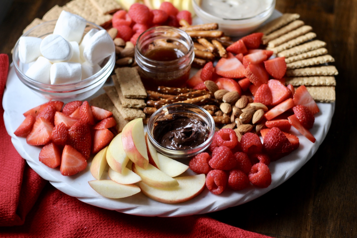 A white marble board with apples, strawberries, raspberries, pretzels, graham crackers, and dips with a red towel in front.