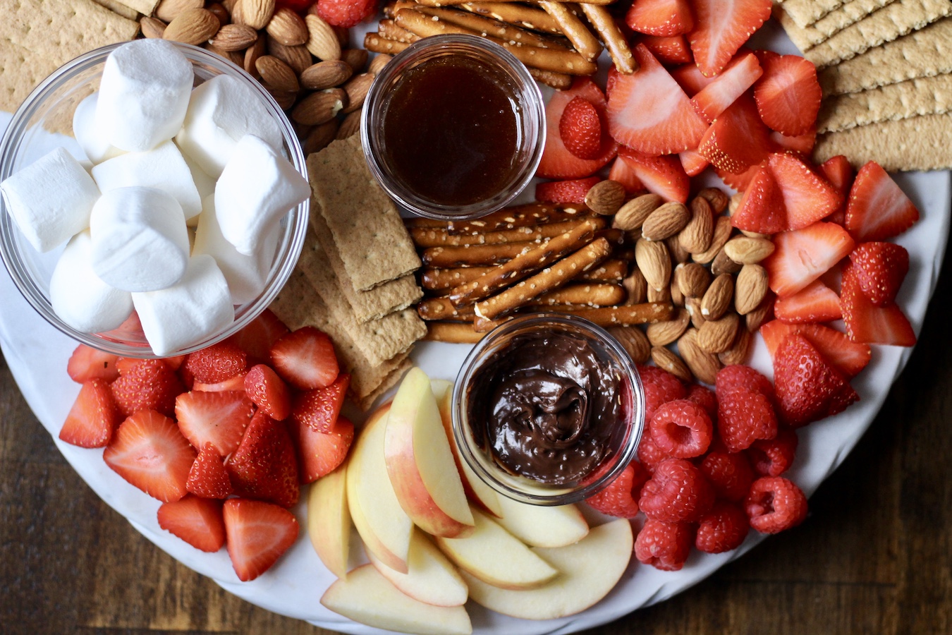 A marble board with marshmallows, strawberries, apples, the hazelnut spread, salted caramel sauce, raspberries, and pretzels.