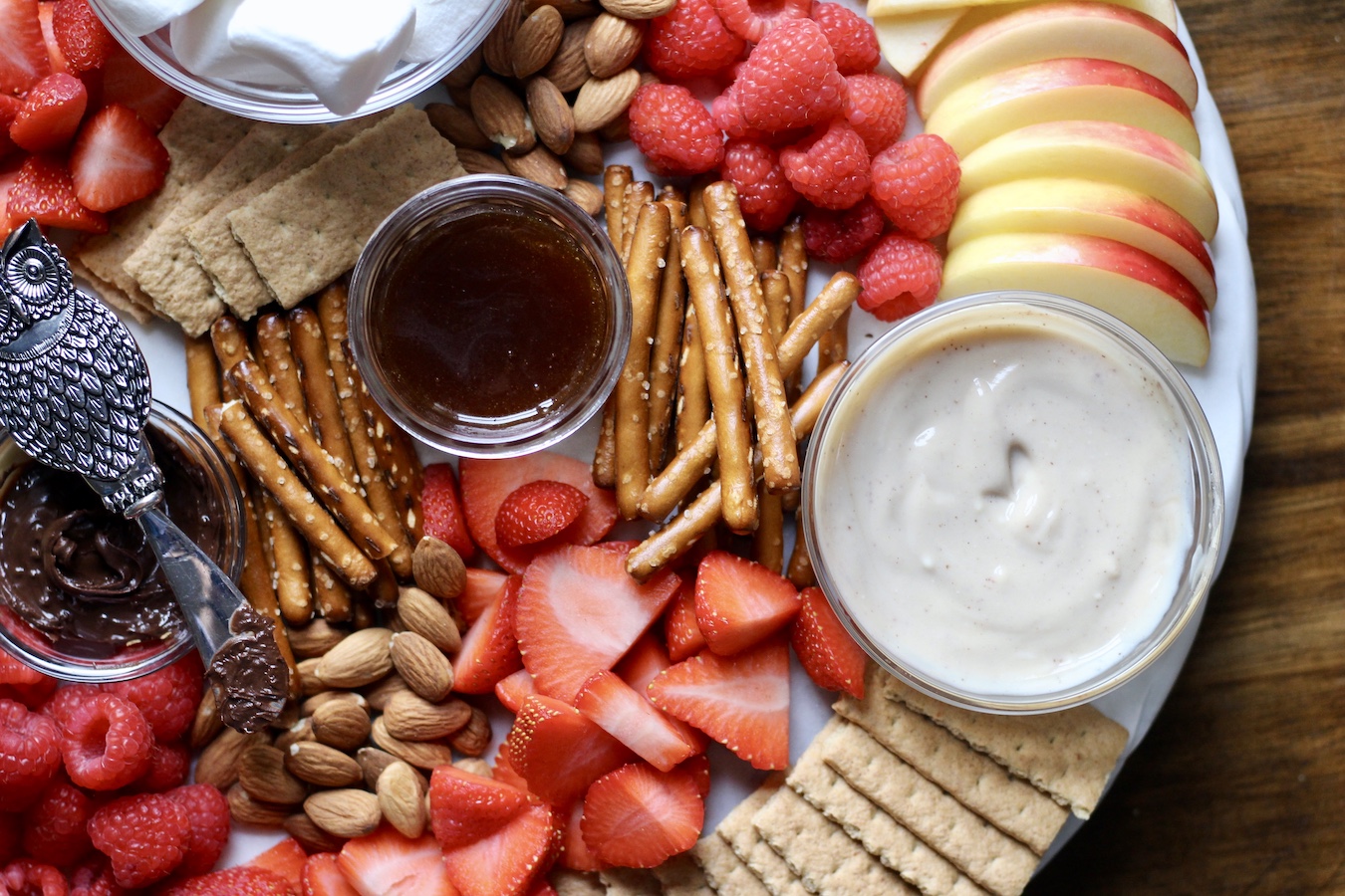The right side of the valentine's day dessert board with yogurt dip, apples, salted caramel sauce, strawberries, pretzels, raspberries, and graham crackers.