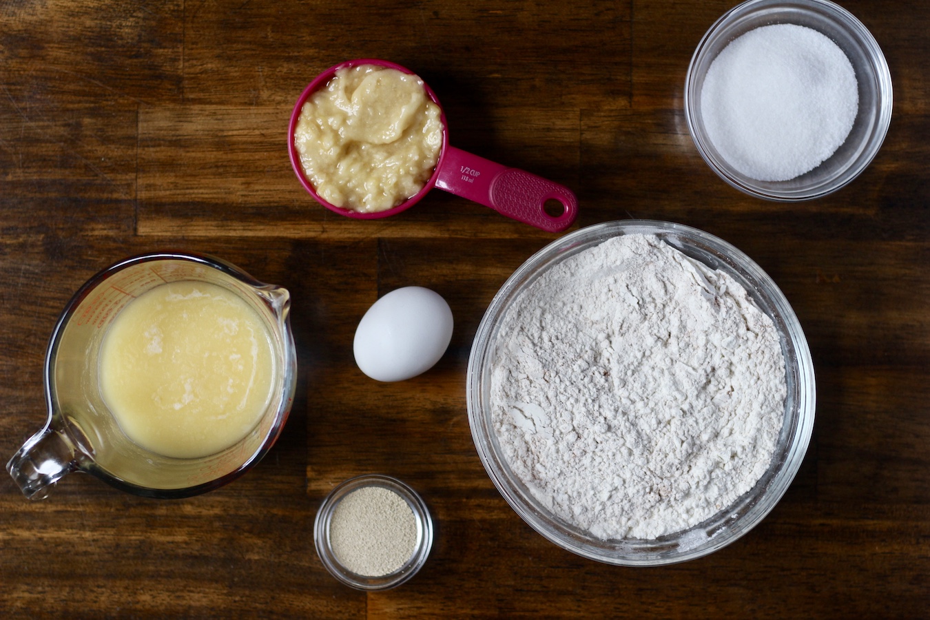 Ingredients to make banana bread cinnamon rolls on measuring cups and glass bowls on a wooden table.