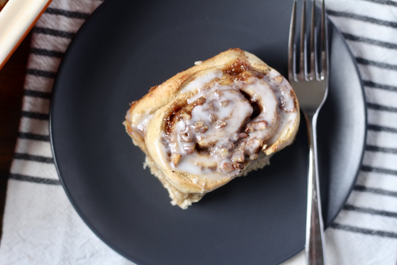 A banana bread cinnamon roll on a blue plate with a fork to the right on a white and blue dish towel.