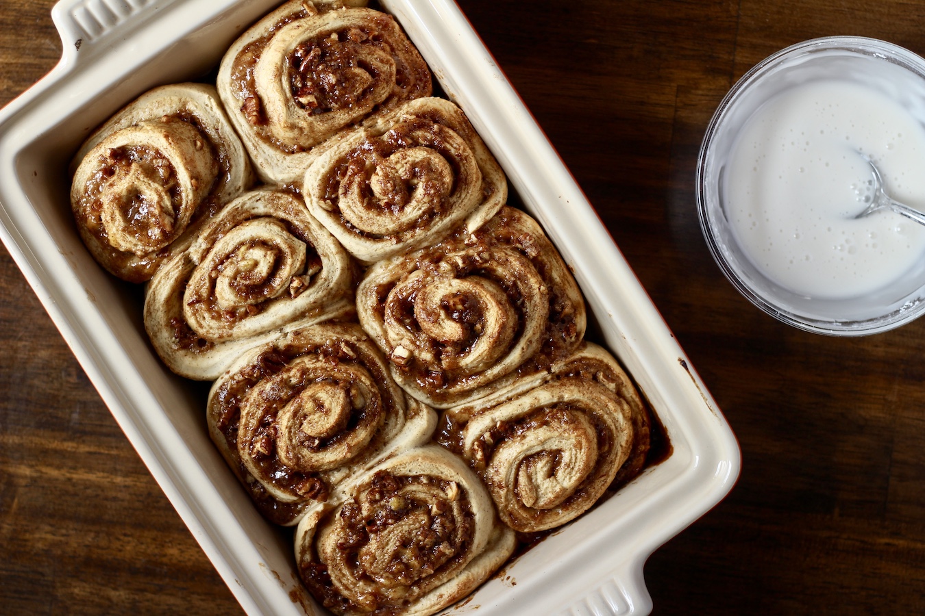 Baked cinnamon rolls on a wooden table with glaze in a glass bowl to the right.