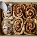 A metal spoon glazing the banana bread cinnamon rolls in the baking dish.