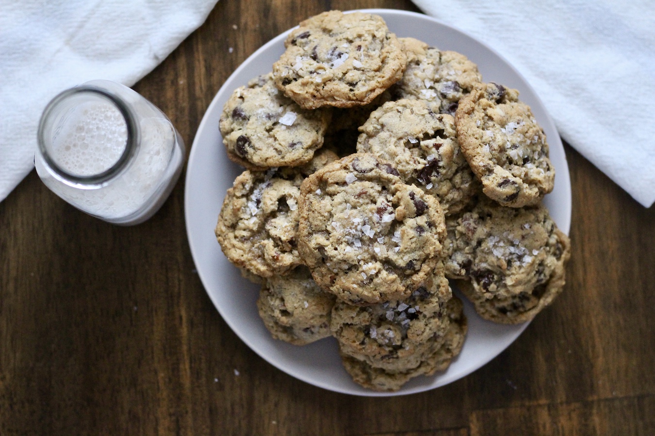 A white plate with a pile of cowboy cookies and a jug of almond milk to the left.
