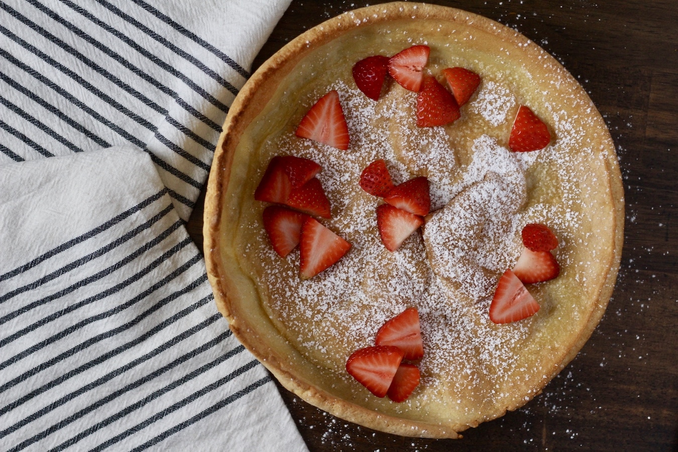 A dutch baby with powdered sugar and sliced strawberries on a plate on a wooden table with a white and blue striped towel to the left.