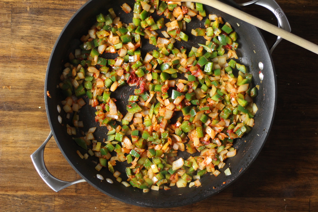 Non-stick skillet with onion, bell pepper, garlic, and tomato paste with a wooden spoon on a wooden table.