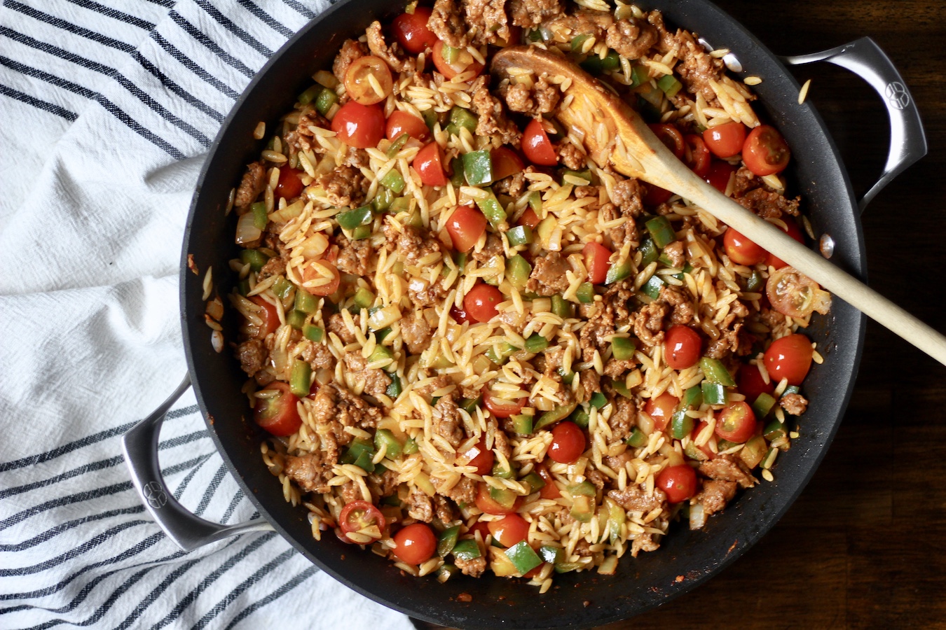 Non-stick skillet with sausage and veggie orzo with a wooden spoon on a wooden table with a white and blue striped towel.