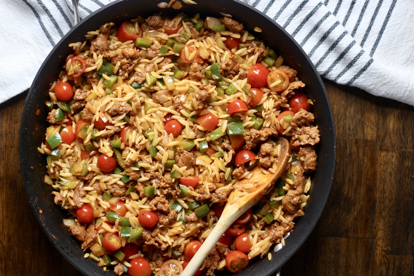Non-stick skillet with sausage and veggie orzo with a wooden spoon on a wooden table with a white and blue striped towel.